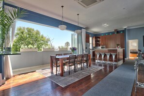 A modern kitchen with a dining area, wooden flooring, and a view of trees outside.