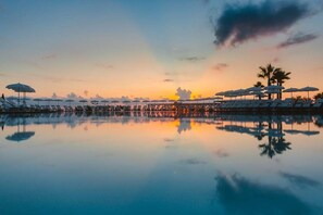 Ein friedlicher Sonnenuntergang über einem ruhigen Pool mit Liegestühlen und Sonnenschirmen, wobei sich Himmel und Palmen spiegeln.