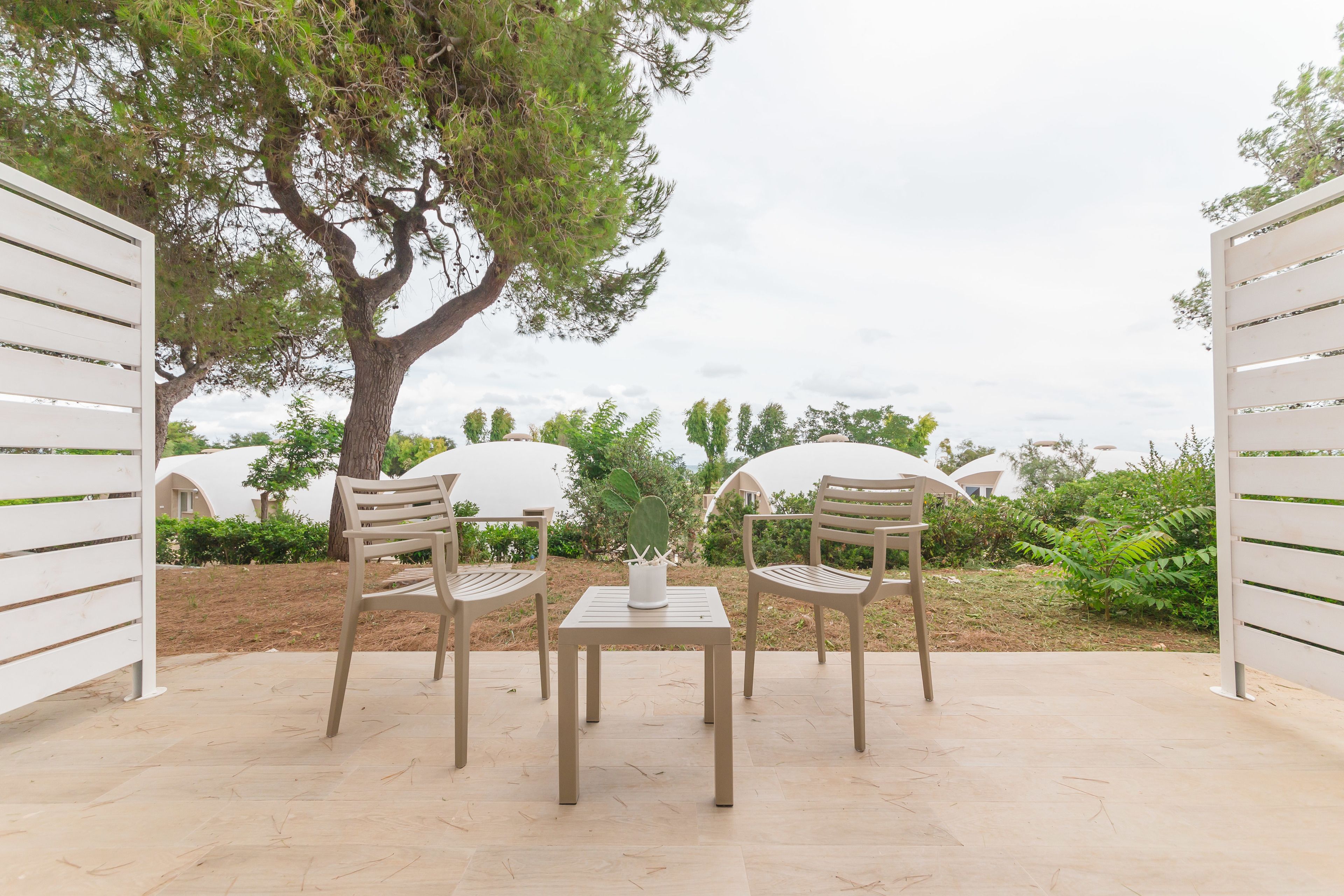 An outdoor seating area with two chairs and a table, surrounded by greenery and a tree.