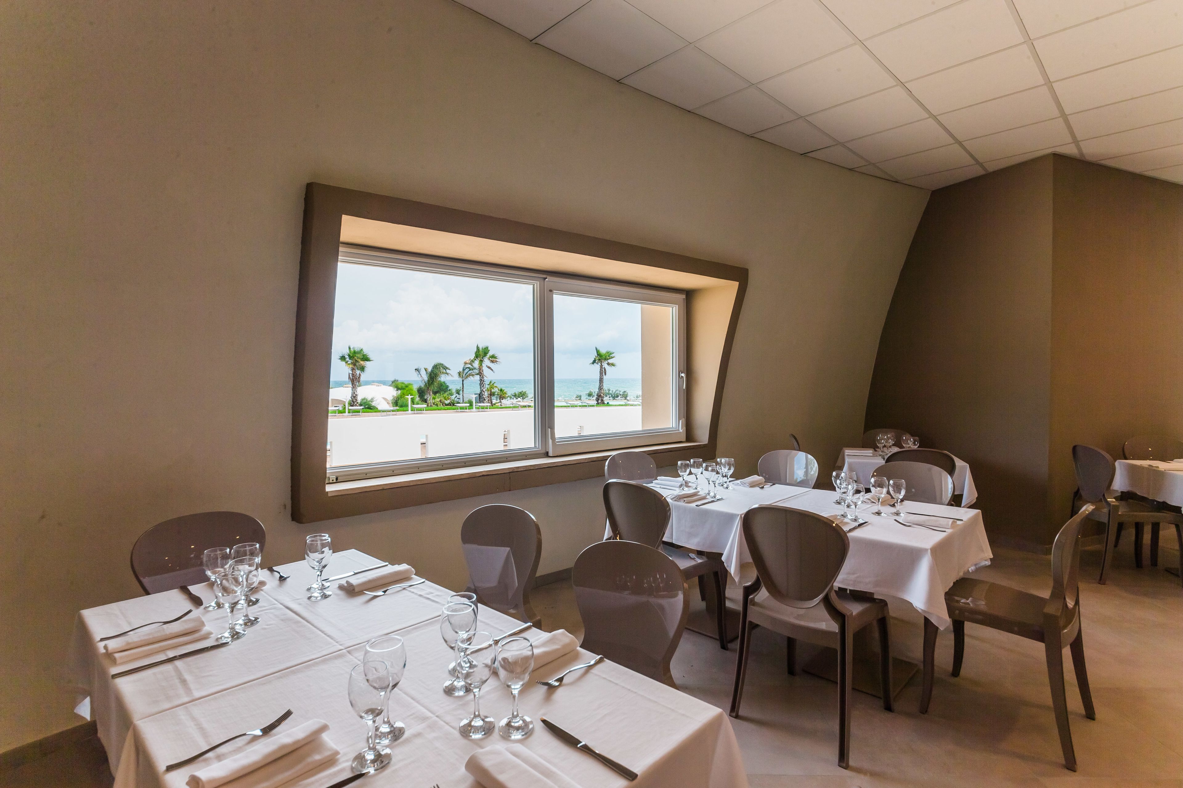 Comedor con mesas dispuestas para el servicio, un ventanal con vistas a palmeras y a la playa, y un techo curvo.