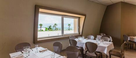 Comedor con mesas dispuestas para el servicio, un ventanal con vistas a palmeras y a la playa, y un techo curvo.