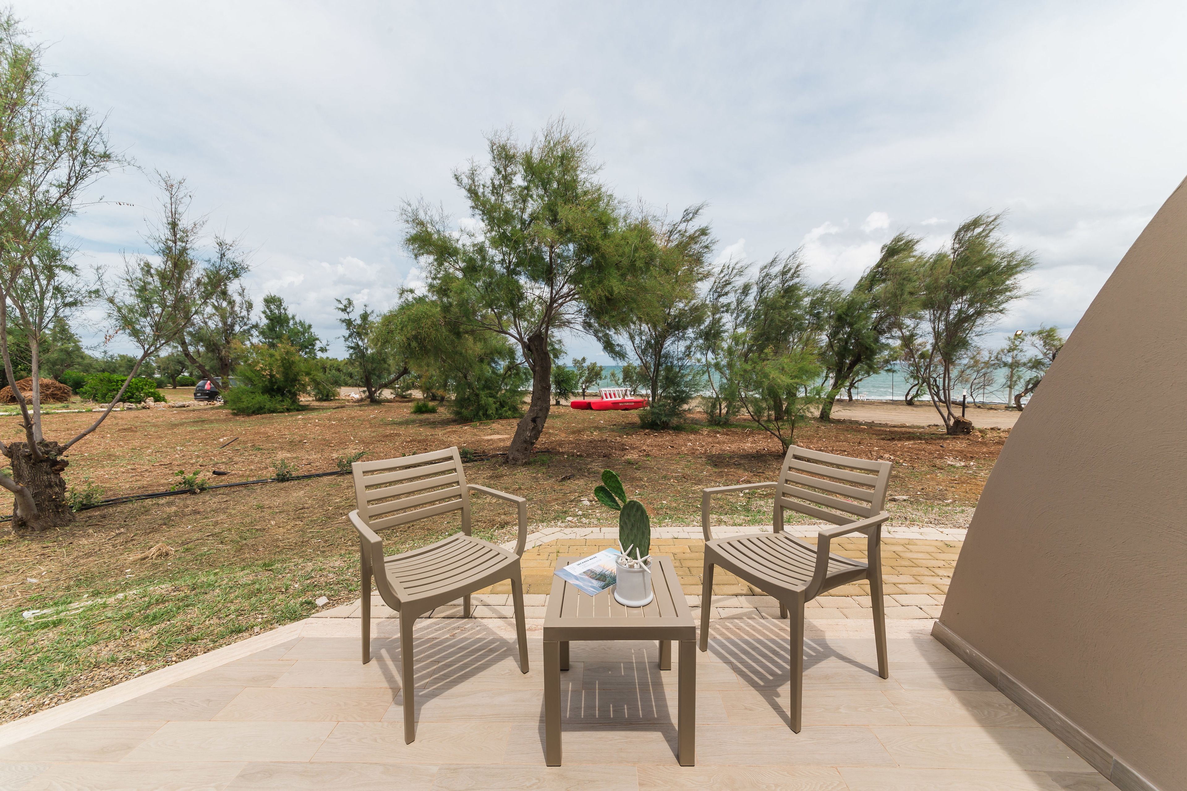 Un espace extérieur aménagé avec deux chaises en bois et une table, entouré d’arbres et offrant une vue sur la mer.