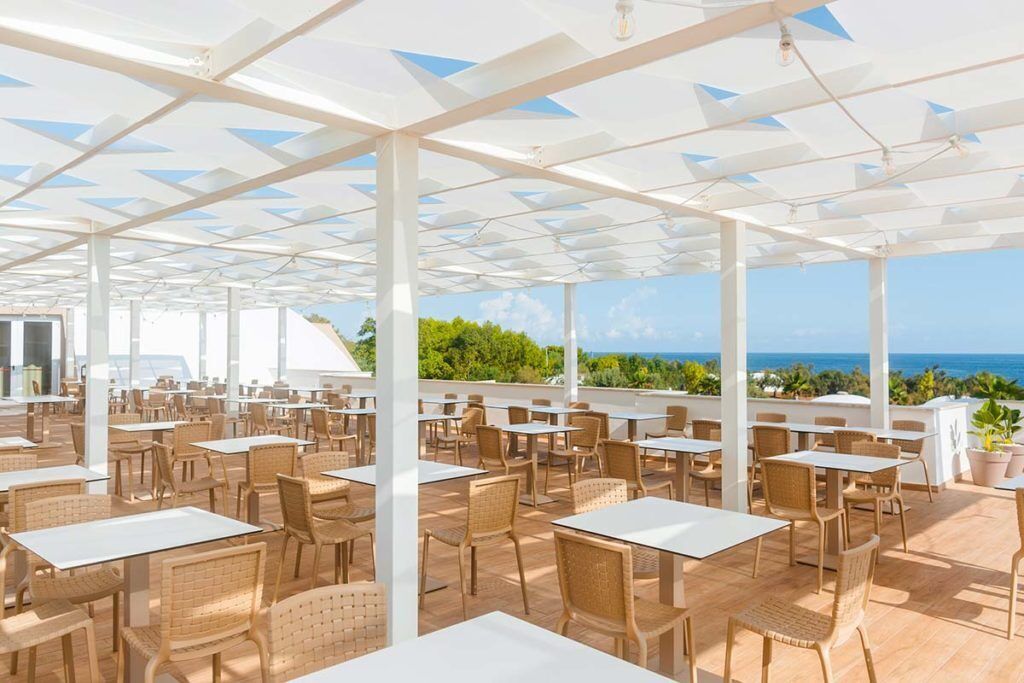 An outdoor dining area with white tables and wicker chairs, under a white pergola with a view of the ocean.