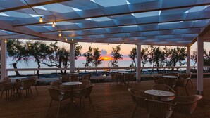 An outdoor seating area with tables and chairs, overlooking a sunset over the water.