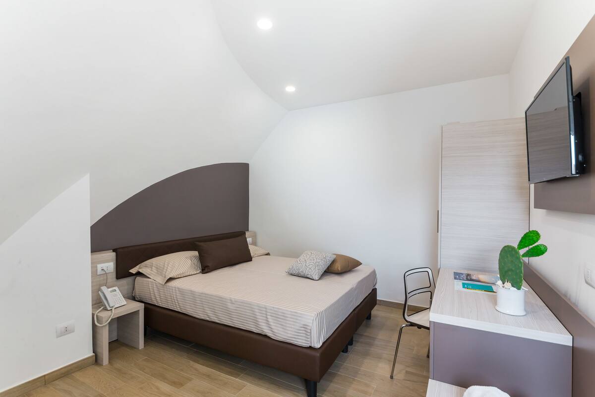 A bedroom with a bed, bedside table, phone, chair, and a cactus on a dresser.