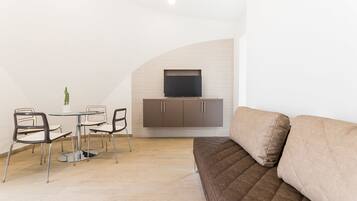 Ein modernes Wohnzimmer mit einem braunen Sofa, einem an der Wand befestigten Fernseher, einem Esstisch mit Stühlen und einer kleinen Topfpflanze.