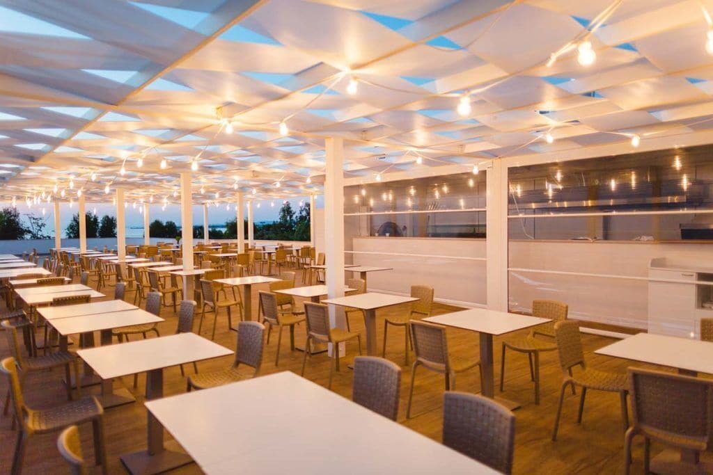 A spacious outdoor dining area with multiple tables and chairs, a modern ceiling design, and a view of the water in the background.