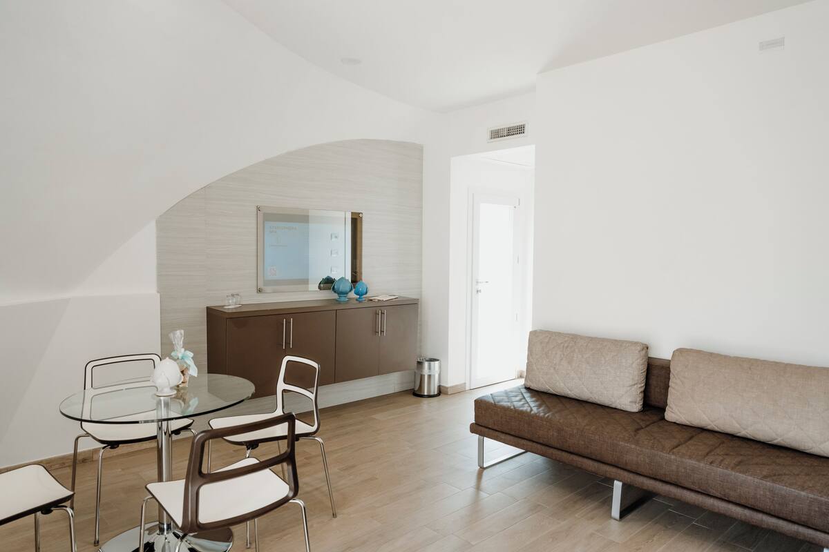 A modern living room with a glass dining table, a brown sofa, and a kitchen area with a white countertop and cabinets.
