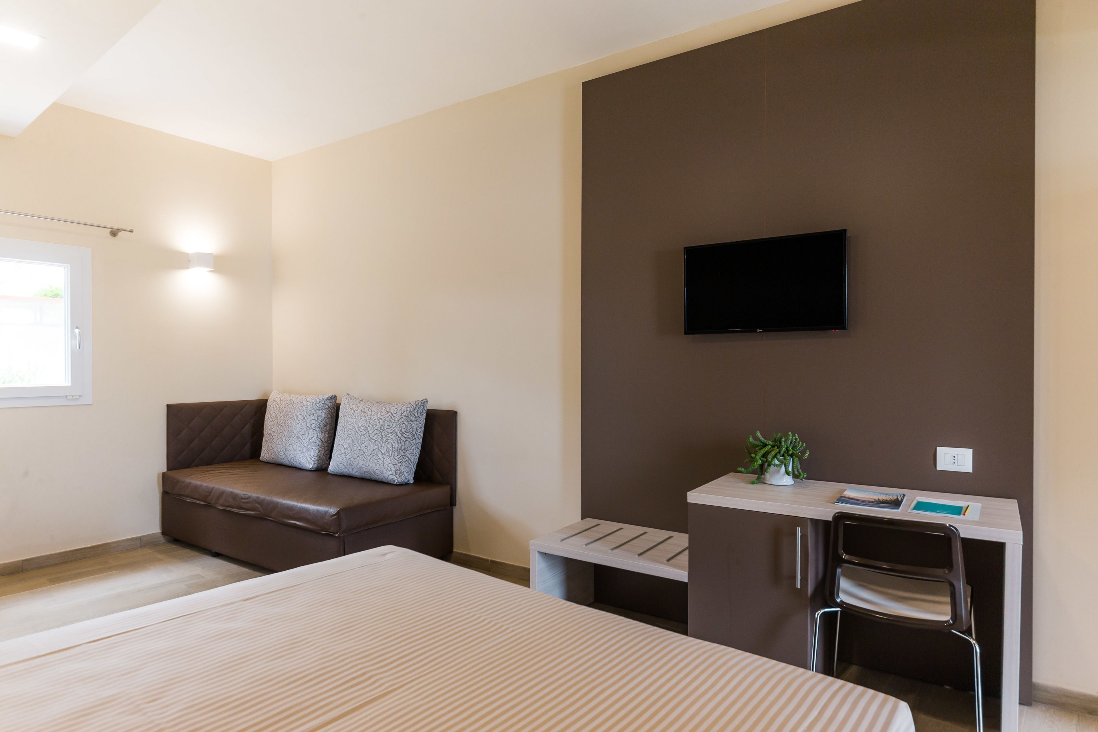 A modern bedroom with a bed, a sofa, a desk with a chair, and a TV mounted on the wall.