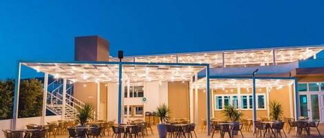 Un espace extérieur aménagé en terrasse avec des tables et des chaises, un coin salon couvert agrémenté de guirlandes lumineuses, et un escalier menant à un niveau supérieur.