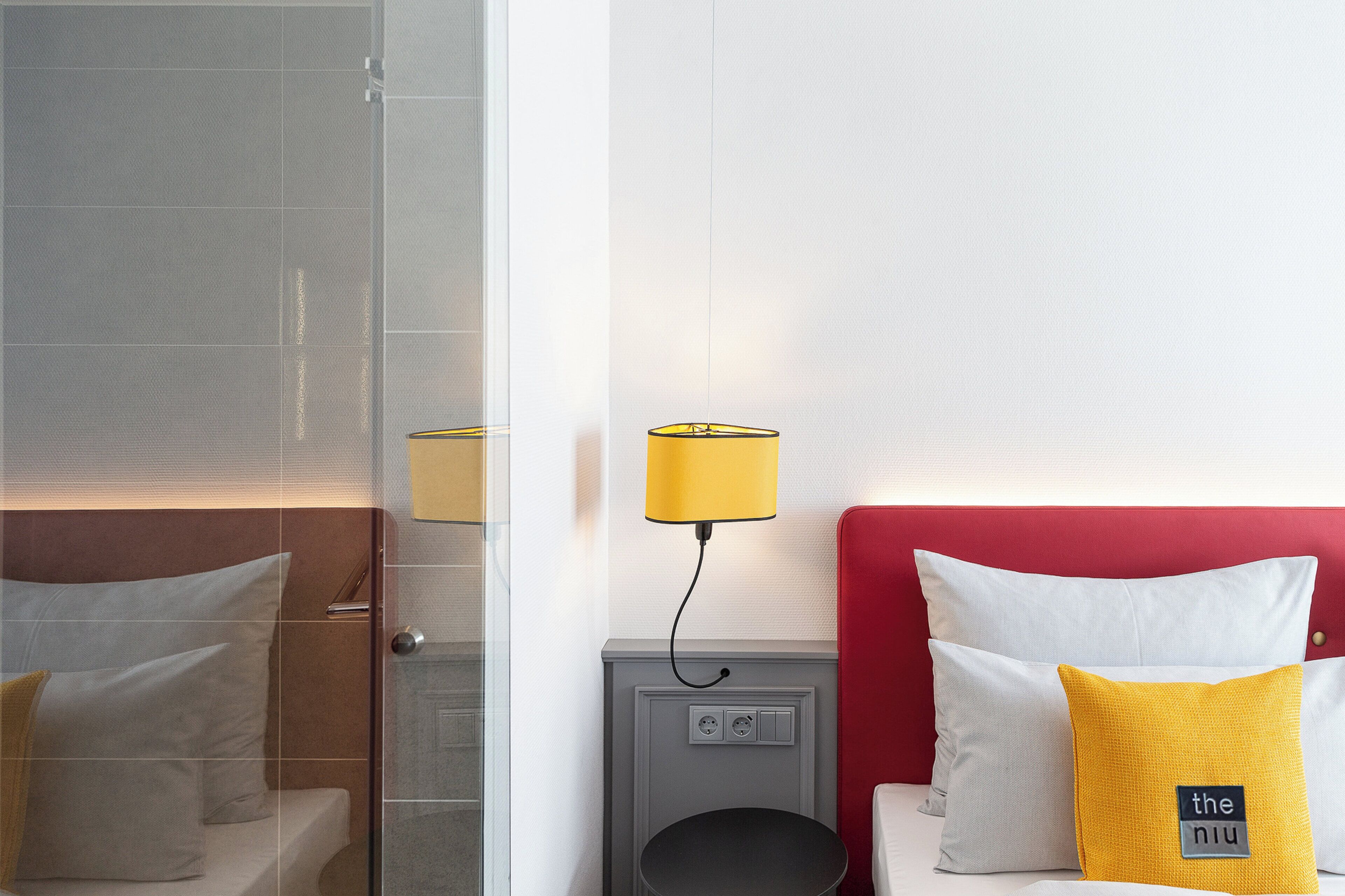 A hotel room with a glass shower enclosure, a bedside table with a lamp, and a red headboard with pillows.