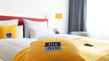 Habitación de hotel con cama, ropa de cama blanca y amarilla, cabecera roja y una mesita de noche con lámpara.