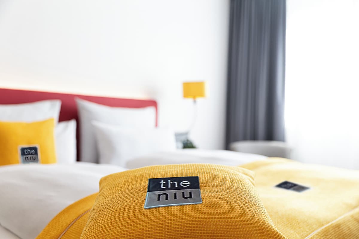 A hotel room with a bed featuring yellow and white bedding, a red headboard, and a small bedside table with a lamp.