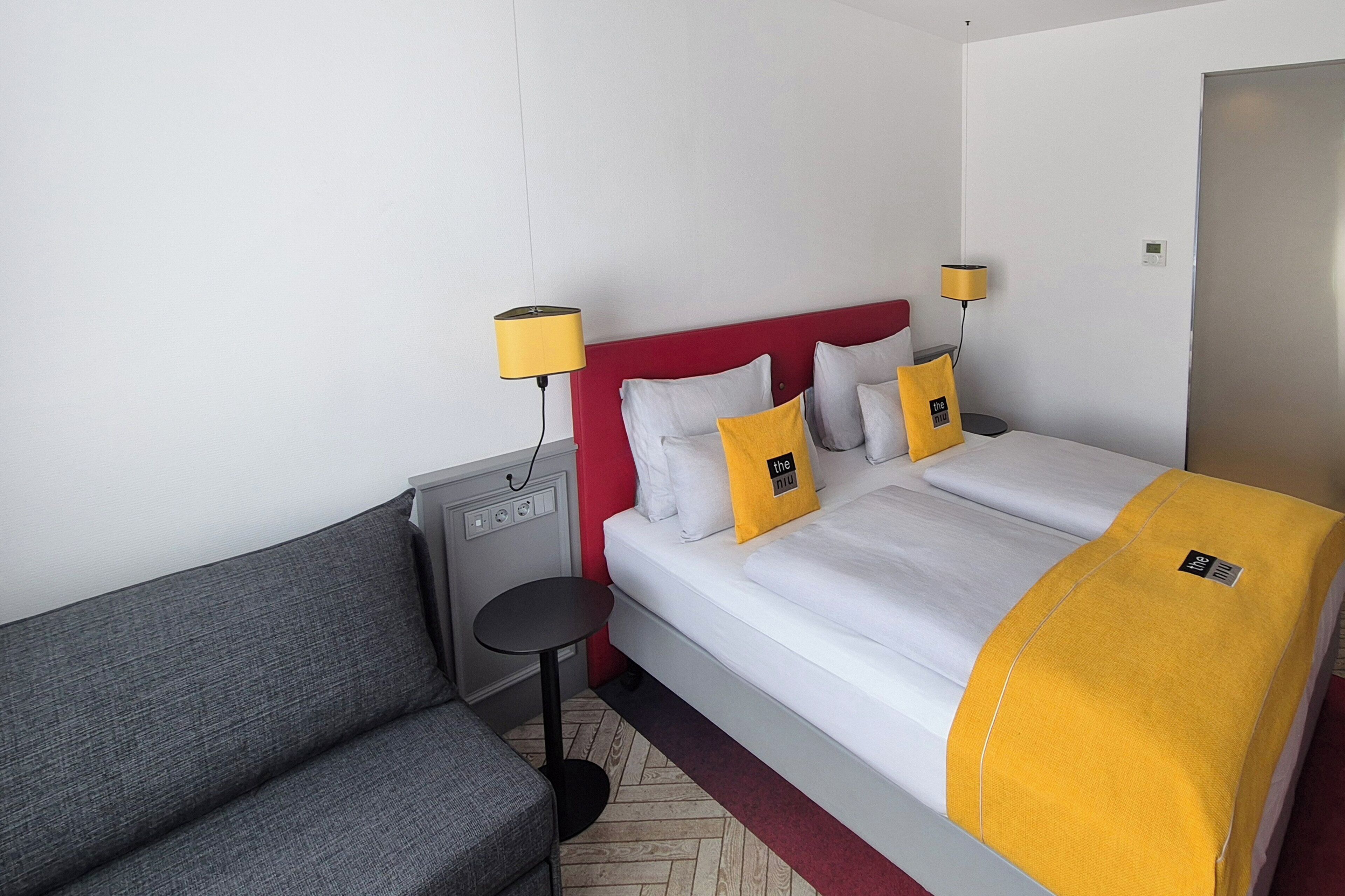 Una habitación de hotel con una cama, un sofá gris, una mesita pequeña y dos lámparas de noche.