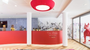 A reception area with a red counter, a receptionist, and a large window with a cityscape view. - Holiday Inn - the niu, Franz Vienna by IHG (Vienna)