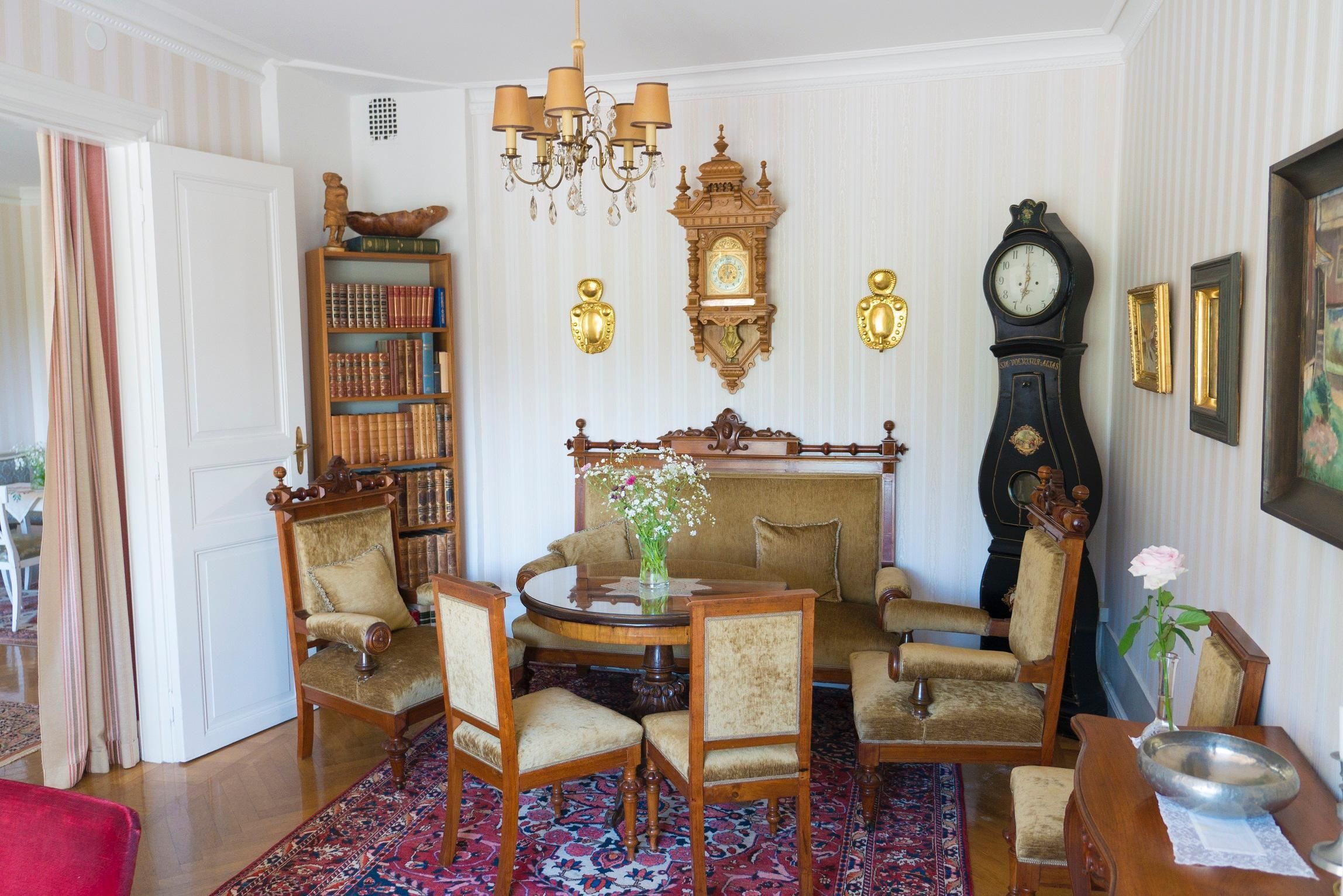 A traditional room with a round table, chairs, a bookshelf, a fireplace, and a grandfather clock.
