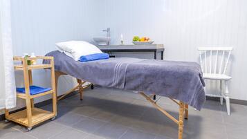 Een kleine, verzorgde kamer met een eenpersoonsbed, een houten plank met toiletartikelen, een witte stoel en een klein tafeltje met een schaal fruit.