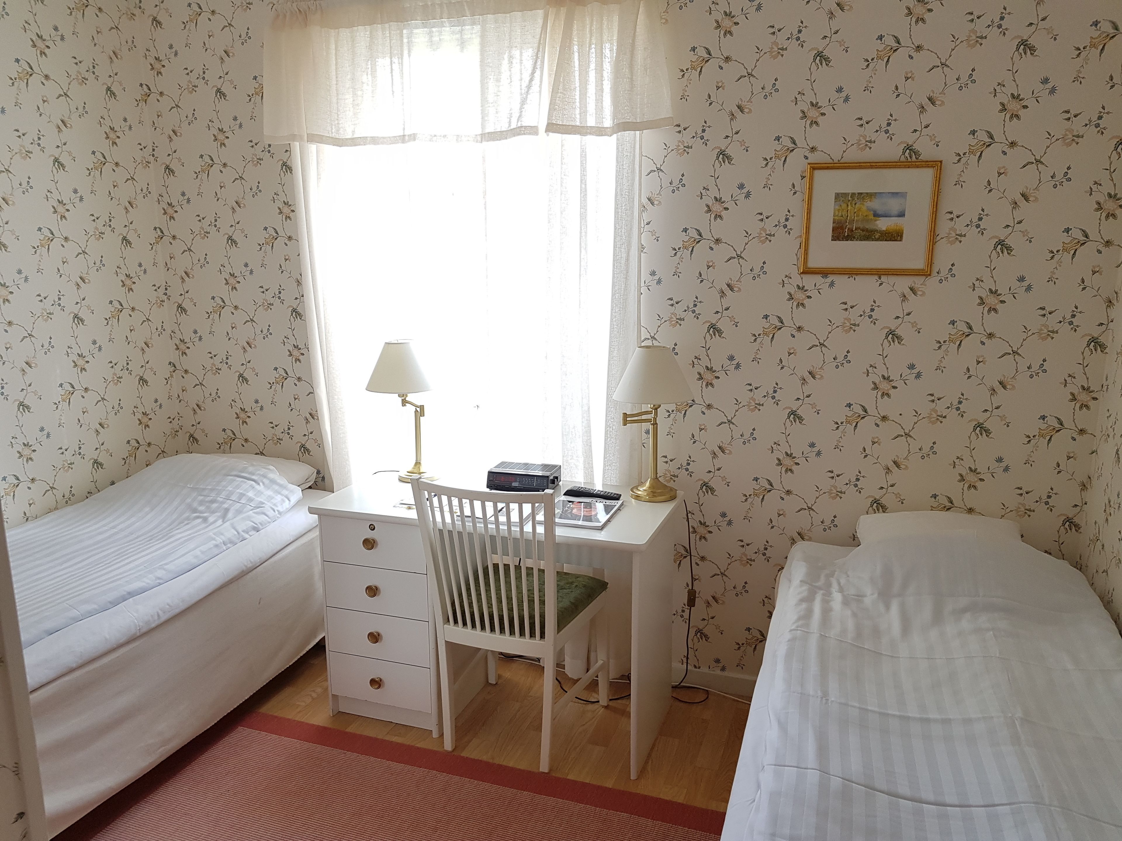 A bedroom with two single beds, a desk with a chair, a lamp, and a framed picture on the wall.