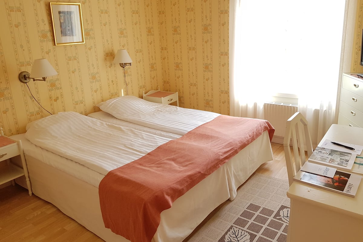 A hotel room with two beds, a desk, a chair, a lamp, and a window with curtains.