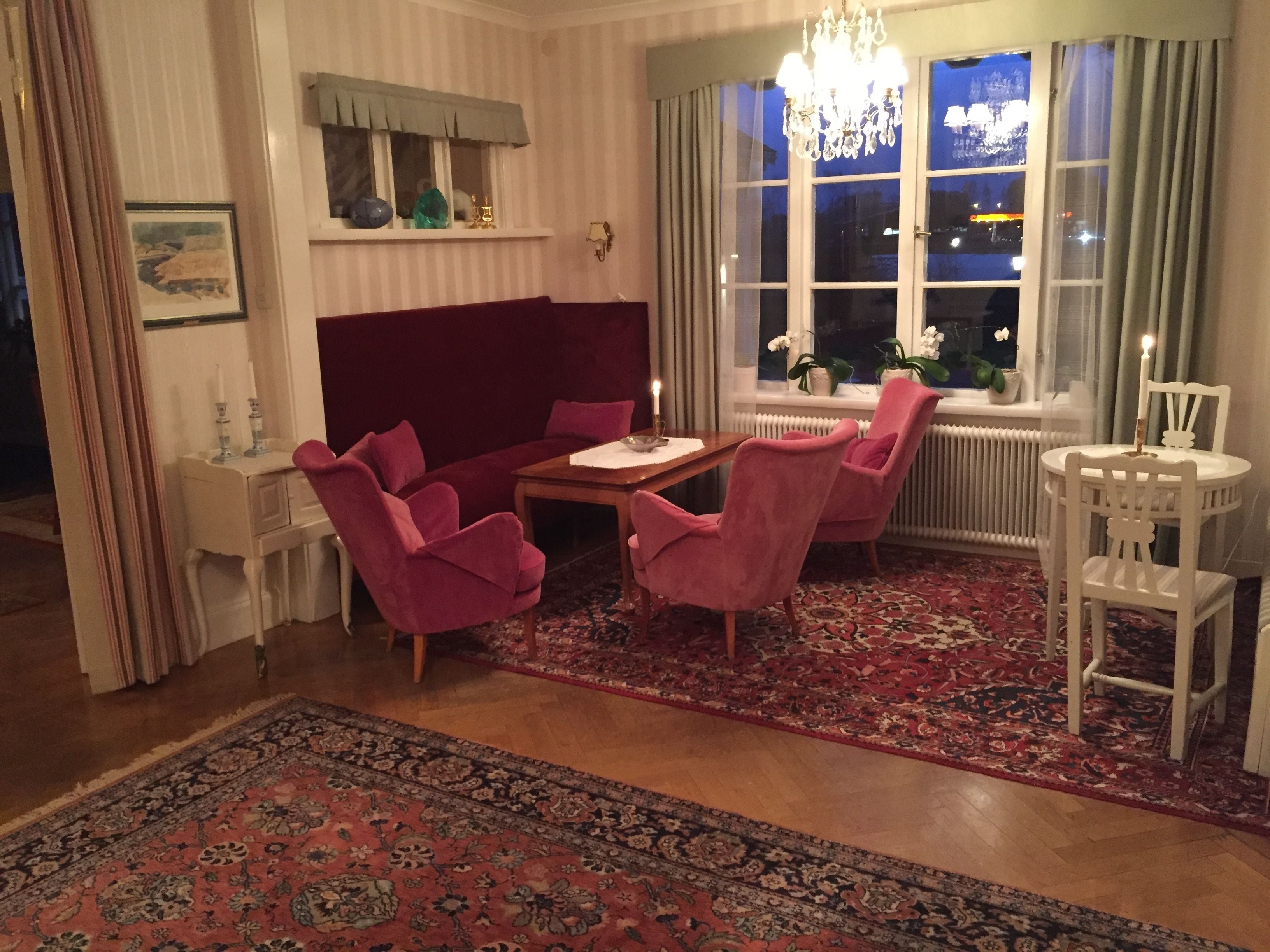 A room with a red sofa, two pink armchairs, a wooden table, and a white chair. There is a window with a view of the night sky and a chandelier.