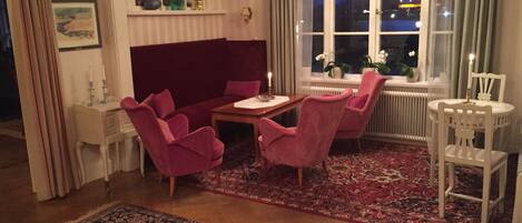 A room with a red sofa, two pink armchairs, a wooden table, and a white chair. There is a window with a view of the night sky and a chandelier.