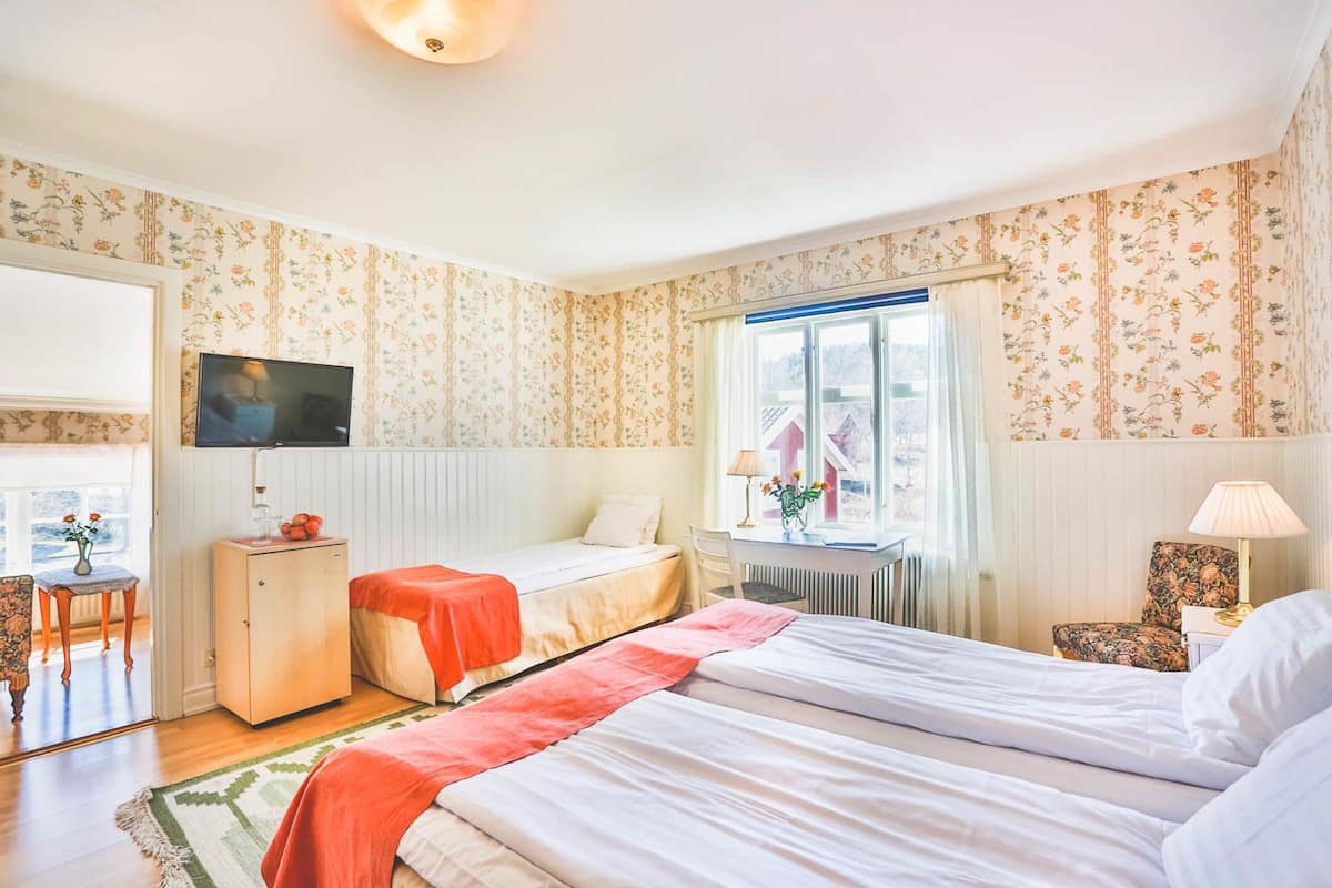 A hotel room with two beds, a TV, a desk, a chair, a window with curtains, and a radiator.
