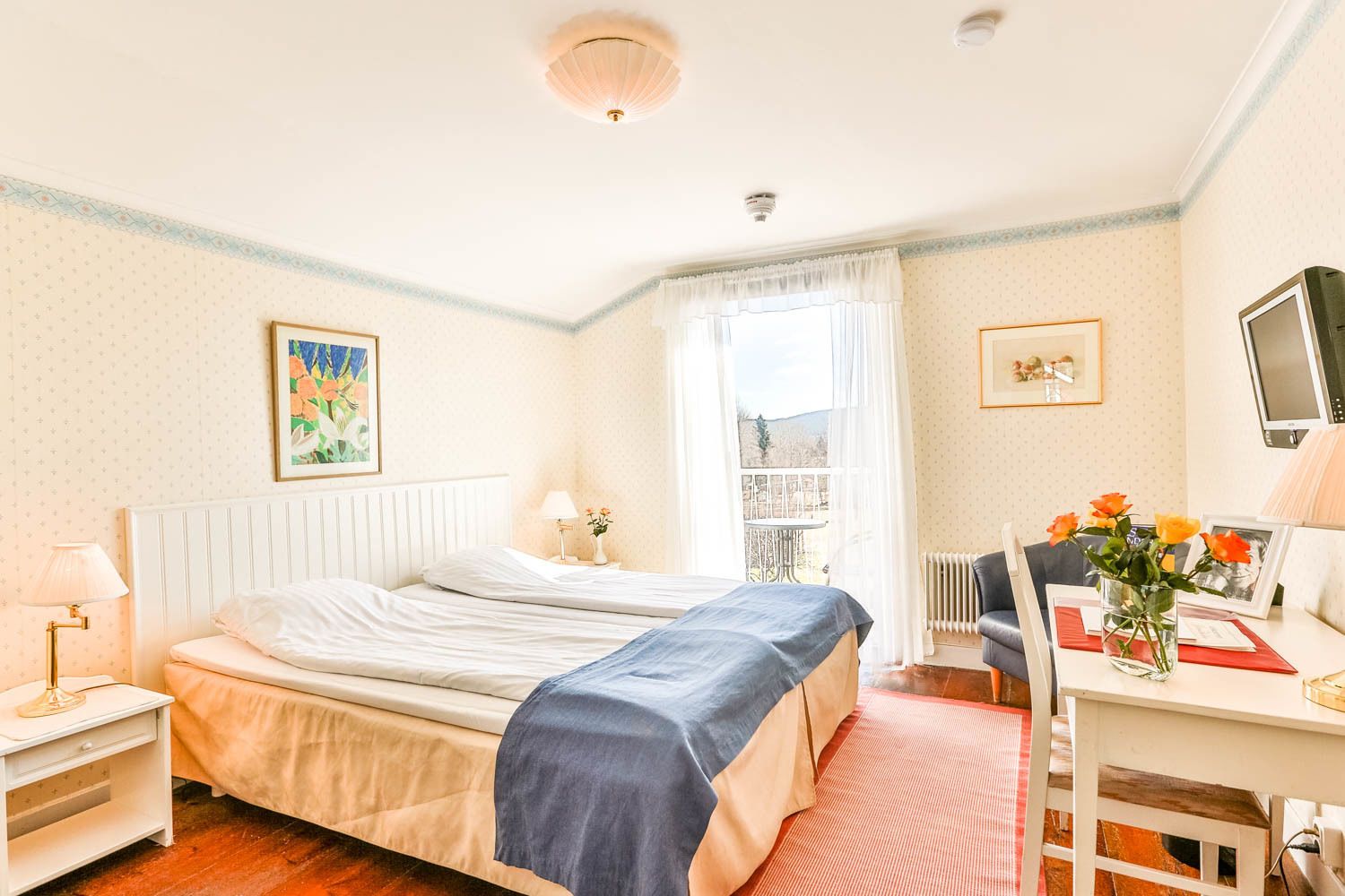 A bedroom with a bed, bedside table, lamp, window with curtains, and a desk with a chair and flowers.