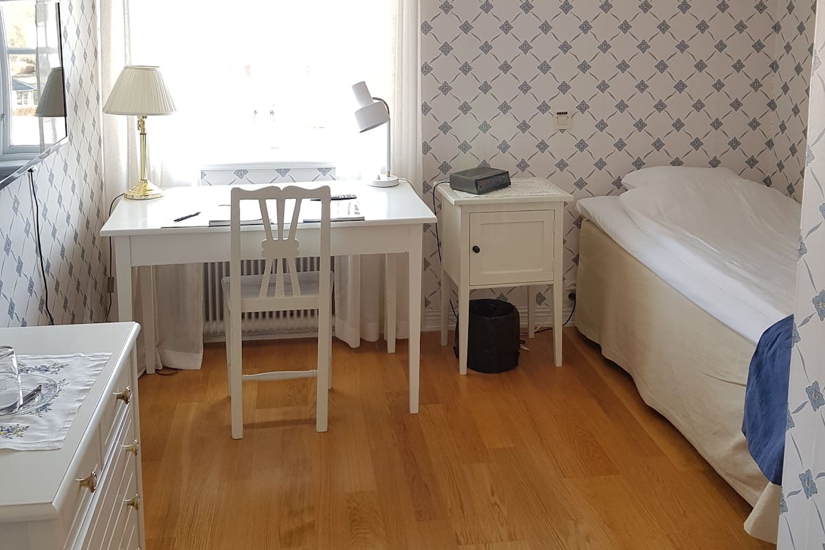 A bedroom with a bed, a desk with a chair, a small bedside table, and a radiator.