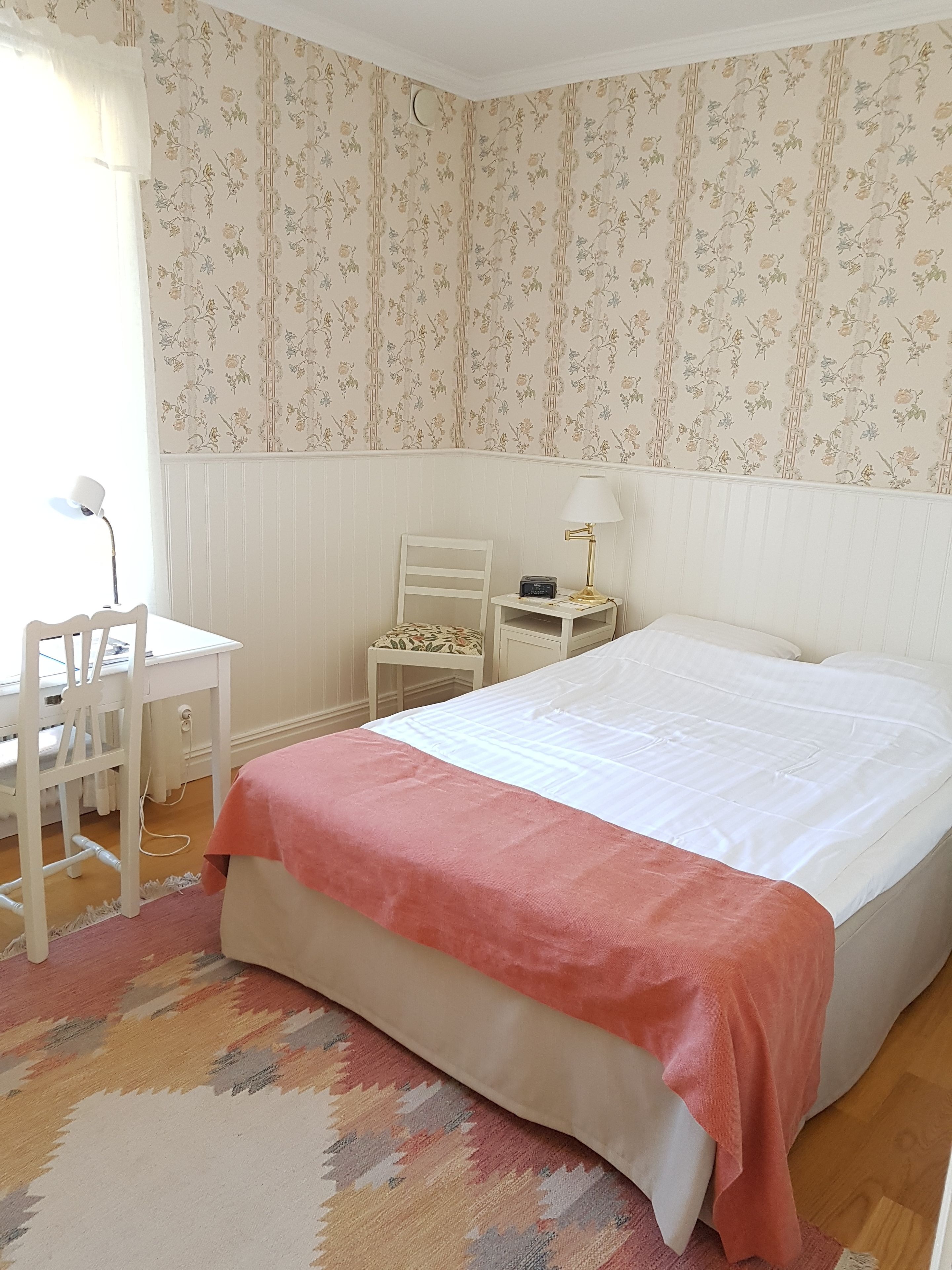 A bedroom with a bed, a desk, a chair, a nightstand, and a lamp.