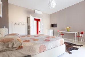 A bedroom with a bed, a desk, two chairs, a TV, and a door with a red frame. - Appartamento Corso Roma (Gallipoli)