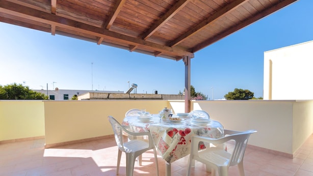 Une terrasse couverte avec une table dressée pour le repas, entourée de chaises, et offrant une vue sur le paysage urbain.