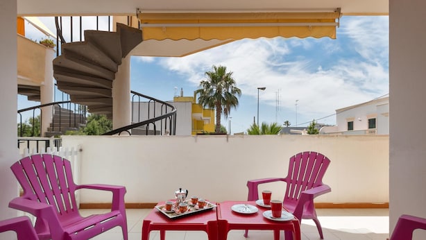 Un balcon agrémenté d’un escalier en colimaçon, deux chaises roses, une table avec un plateau de tasses et une théière, et une vue sur des immeubles et un palmier.