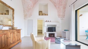 Un salón amplio con un armario de madera, un sillón color crema, una mesa de centro de vidrio y un televisor sobre un soporte blanco.