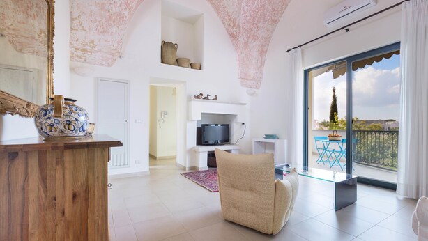 Un salón espacioso con una mesa de centro de madera, un sillón beige y un televisor. Hay un balcón con vistas a la ciudad.
