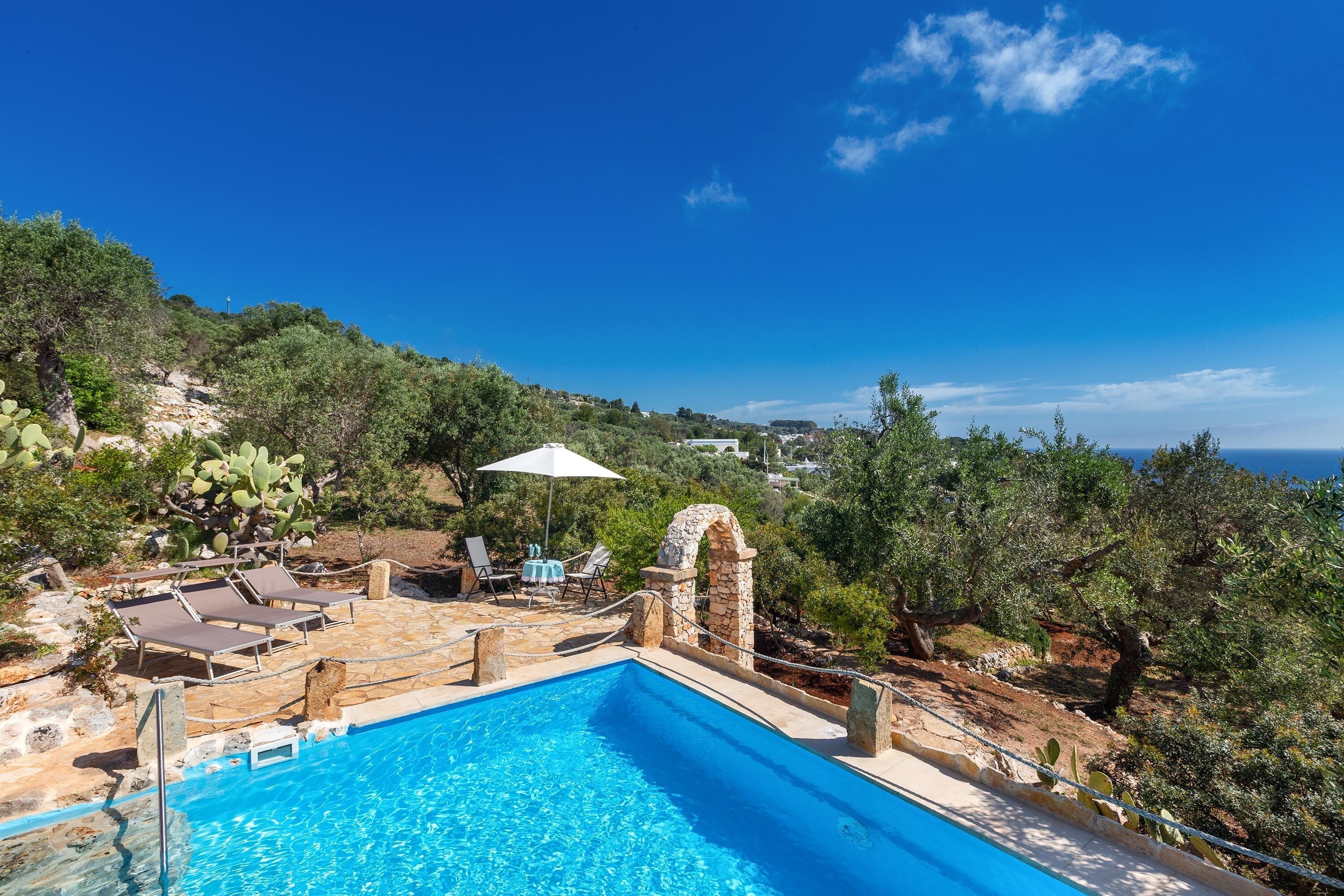 A pool with lounge chairs and a stone archway, surrounded by greenery and overlooking a coastal area.