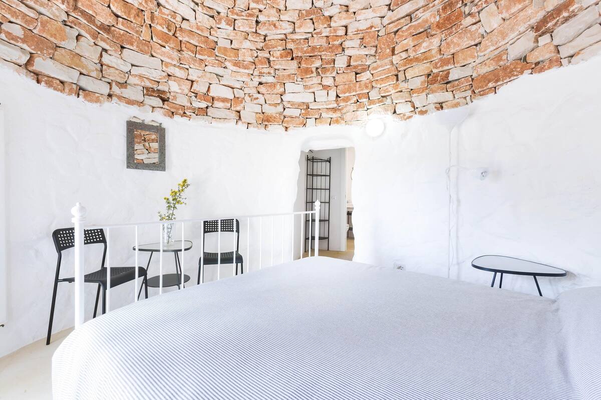 Ein Zimmer mit Ziegelmuster-Decke, weißen Wänden, einem Bett mit gestreifter Bettdecke, zwei schwarzen Stühlen, einem kleinen runden Tisch und einer Topfpflanze.