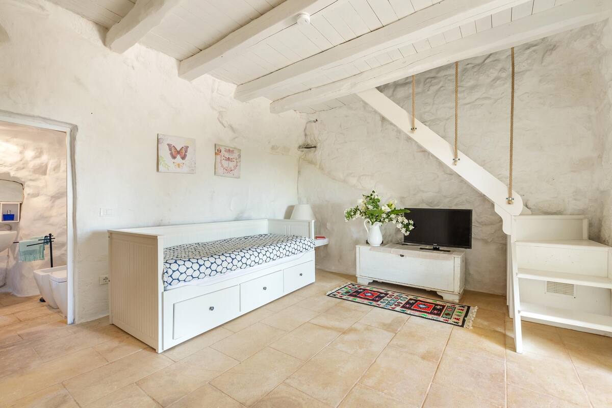 Ein Zimmer mit einem weißen Bett, einem kleinen weißen TV-Schrank mit Fernseher, einem weißen Schrank, einer weißen Bank, einem weißen Regal und einer weißen Treppe.