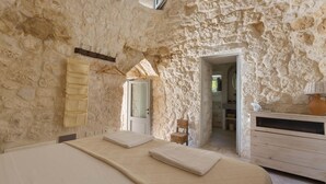 A room with a stone wall, a bed with a textured blanket, a wicker chair, and a wooden dresser. - La Dialma (Ostuni)