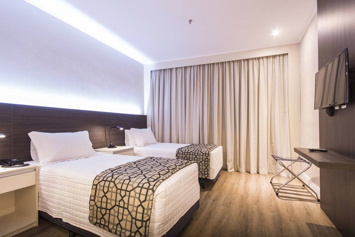 Ein Hotelzimmer mit zwei Betten, einem Schreibtisch mit Telefon, einem an der Wand montierten Fernseher und einem Fenster mit Vorhängen.