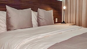 Een opgemaakt bed met een houten hoofdbord, een bedlampje en uitzicht buiten door een raam met gordijnen.