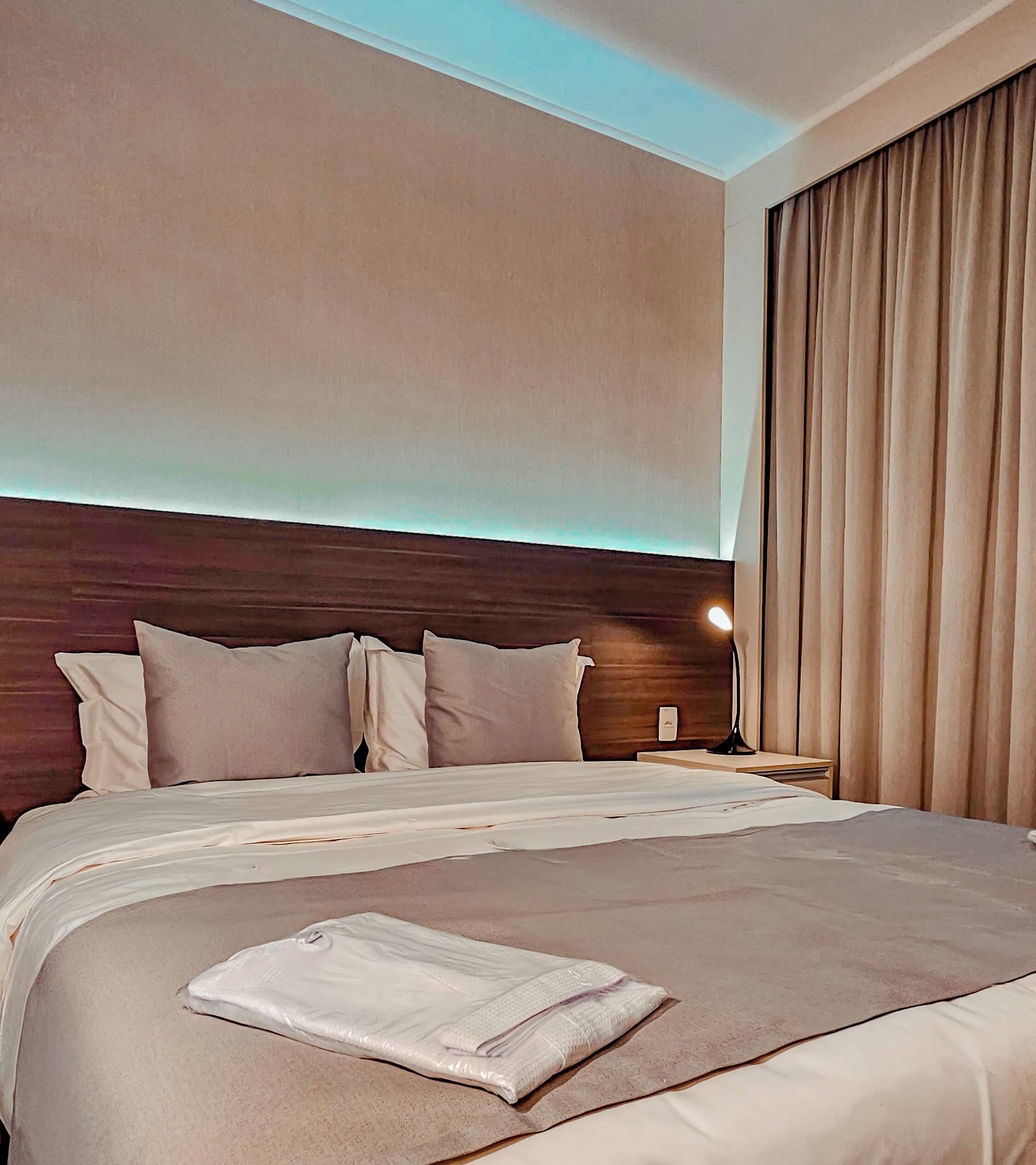 A hotel room with a large bed, a wooden headboard, a nightstand with a lamp, and a window with curtains.