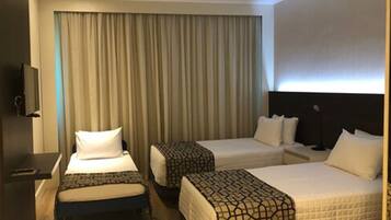 A hotel room with two single beds, a desk with a chair, a TV, and a large window with curtains.