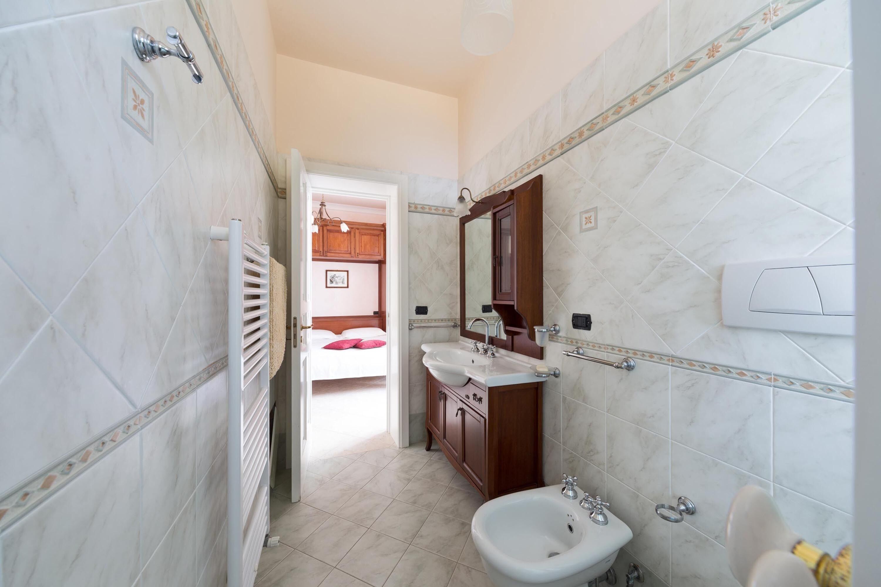 Ein Badezimmer mit Bidet, Waschbecken und Spiegel. Eine Tür führt in einen Schlafbereich mit Bett und einer Pendelleuchte.