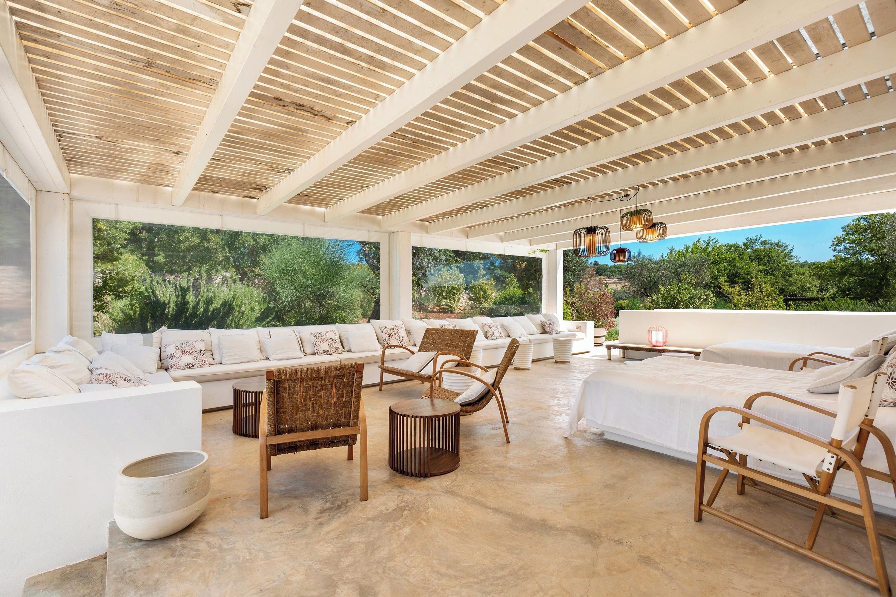 A spacious outdoor lounge area with a wooden ceiling, white furniture, and large windows offering a view of greenery.