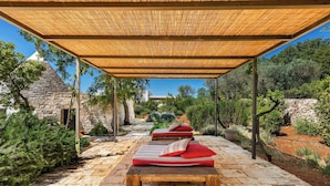 A covered outdoor seating area with wooden benches and red cushions, surrounded by stone walls and greenery. - Le Ali Bianche (Ostuni)
