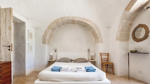 A bedroom with a large bed, a wooden dresser, a chair, a small table with a bowl, and an arched stone ceiling. - Le Ali Bianche (Ostuni)