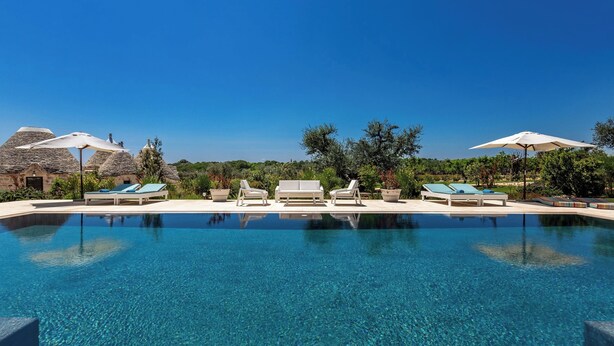 A clear blue infinity pool with lounge chairs and umbrellas, surrounded by greenery and a clear sky. - Le Ali Bianche (Ostuni)
