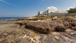 A coastal building with a flat roof and a balcony, situated on rocky terrain with a clear view of the sea. - Lo Scoglio Bianco (Ostuni)