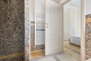Ein Badezimmer mit Duschkabine aus Mosaikfliesen, einer weißen Tür und einem angrenzenden Raum mit einem Bett mit weißer Bettwäsche.