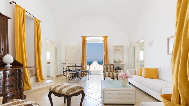A spacious living room with a white sofa, a patterned ottoman, and a coffee table. There is a wooden cabinet and a dining table with chairs. The room has yellow curtains and a view of the sea through an open doorway. - Lo Scoglio Bianco (Ostuni)